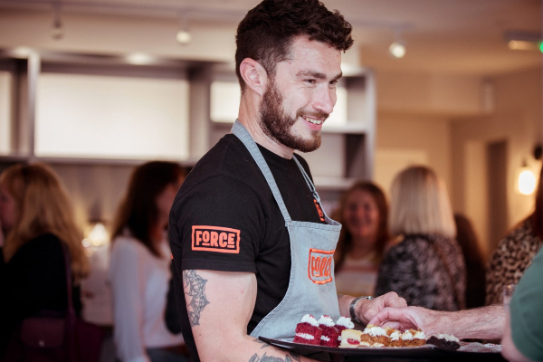 Man in a black 'FORCE' t-shirt and apron holding a plate of food in a social setting.